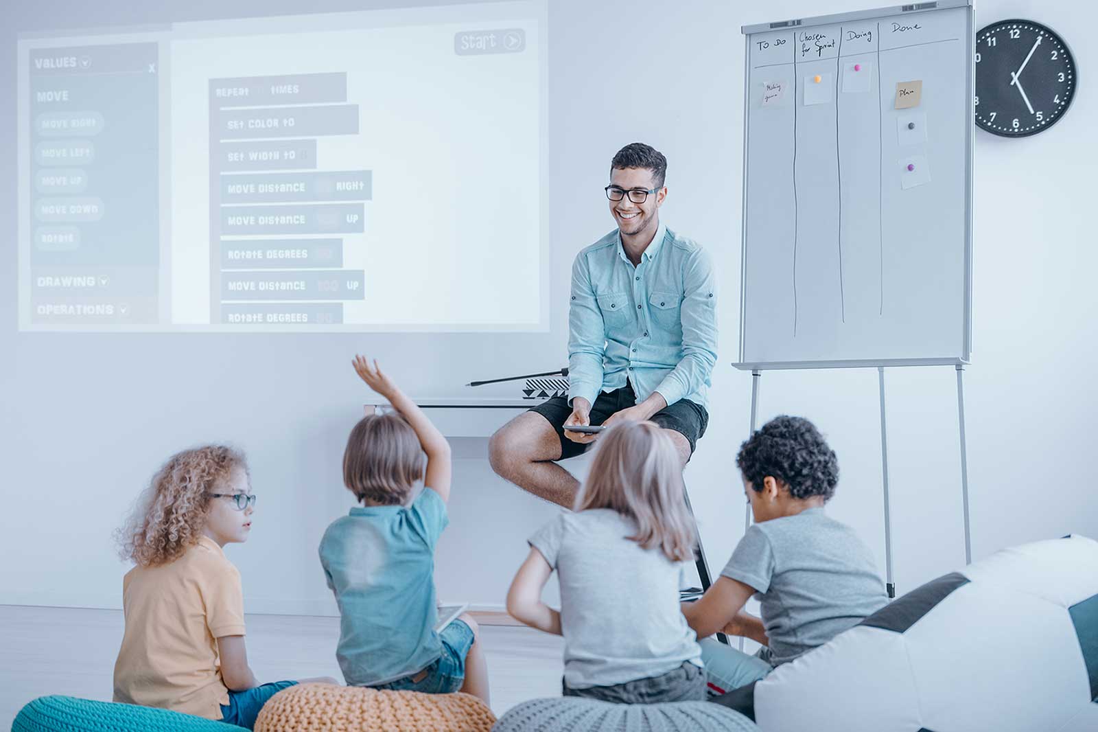 Ein Lehrer sitzt auf einem Hocker, umgeben von engagierten Schülern in einem Klassenzimmer, mit einem Projektor, der Programmierbefehle anzeigt.