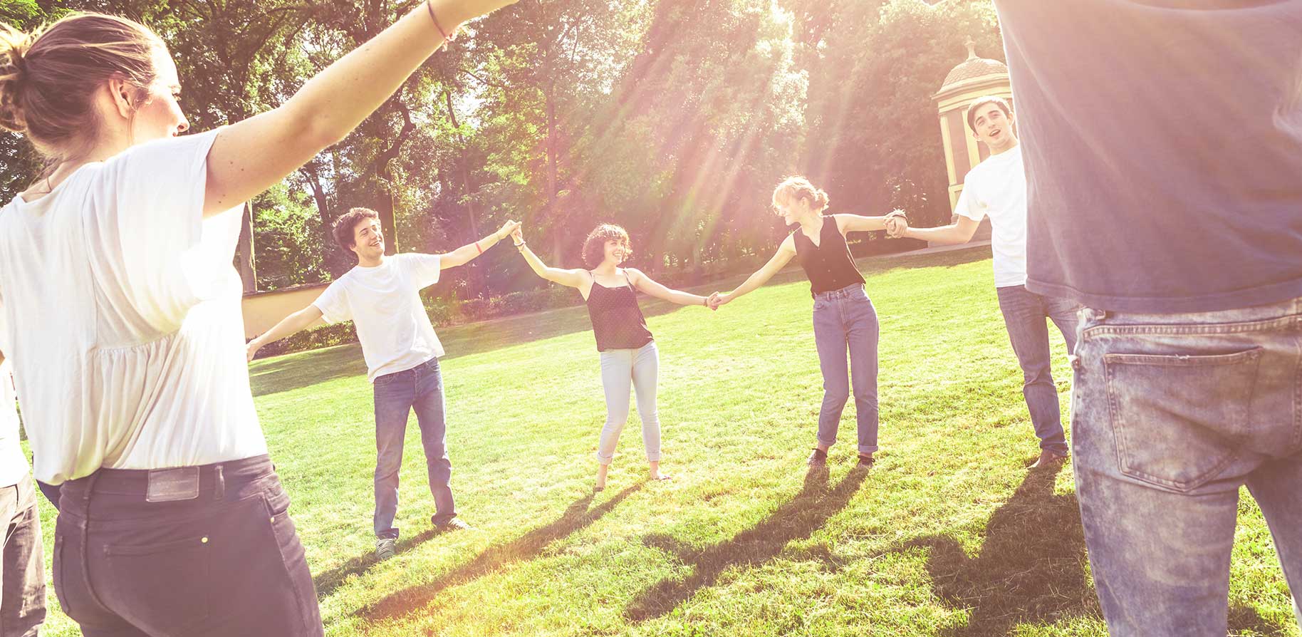 A group of people holds hands in a sunny park. They stand in a circle and appear to be joyful.