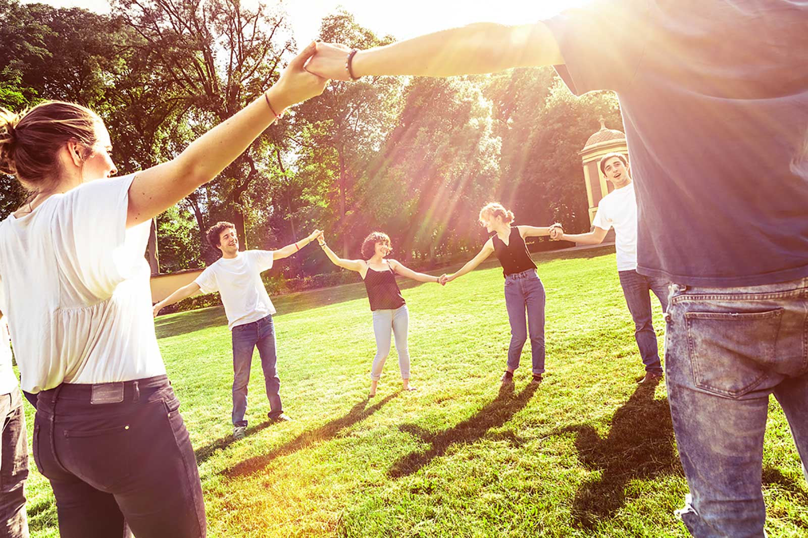 Eine Gruppe von Menschen bildet einen Kreis im Park, die Hände haltend, während die Sonne hinter ihnen scheint.